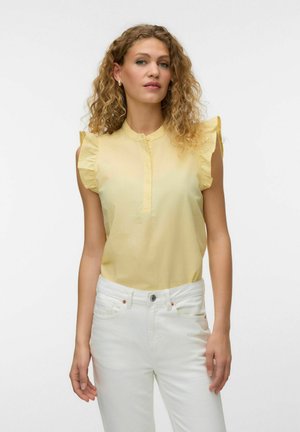 Kobieta o kręconych włosach, ubrana w żółtą, bezrękawnikową bluzkę z falbankami na ramionach i białe spodnie, stojąca na jednolitym tle.