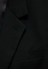 Black suit jacket detail showing a textured fabric, with a lapel and a single pocket. The design features clean lines and a tailored fit.