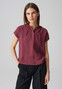 Top corto a maniche corte di colore marrone scuro con cappuccio, presenta una patta con bottoni e una texture morbida, abbinato a pantaloni neri larghi.