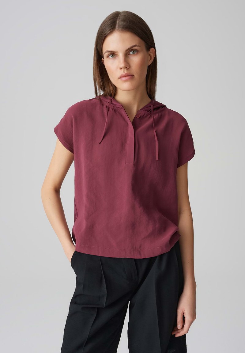 Top corto a maniche corte di colore marrone scuro con cappuccio, presenta una patta con bottoni e una texture morbida, abbinato a pantaloni neri larghi.