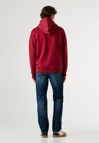 Raudona gobtuvinė džemperis su tekstūruotu paviršiumi, su kengūros kišene. Dėvima su mėlynomis džinsinėmis kelnėmis ir neutralios spalvos batais.