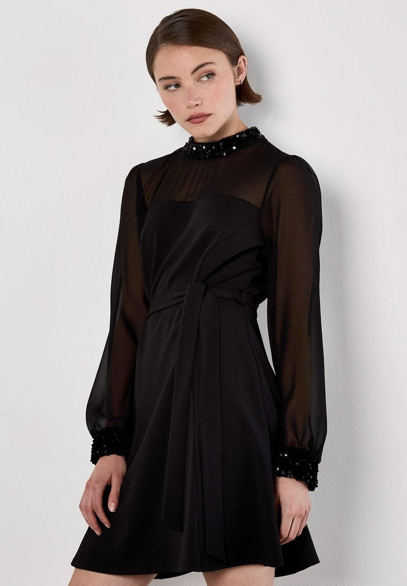 Vestido negro con mangas largas transparentes y un cuerpo ajustado. Presenta un lazo en la cintura y puños adornados con lentejuelas. Textura suave y silueta fluida.