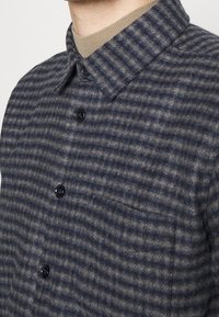 Camicia a quadri in lana di colore blu navy e beige, con colletto, quattro bottoni neri e taschino sul petto. Tessuto testurizzato, con strisce orizzontali.