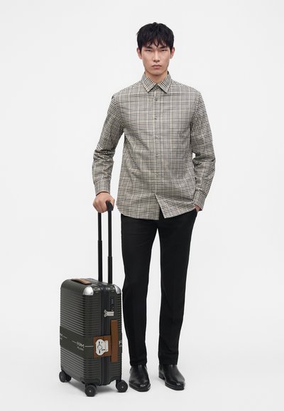 Jeune homme portant une chemise à carreaux et un pantalon noir, debout et tenant la poignée d'une valise noire à roulettes.