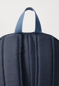 Mørkeblå ryggsekk med polstrede stropper, et håndtak på toppen og et teksturert stoff med sømmer som går langs baksiden for ergonomisk støtte.