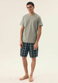 Jasnozielona bawełniana koszulka z kieszonką na piersi, zestawiona z granatowo-niebieskimi szortami w kratę z zielonymi i białymi akcentami. Model jest boso.