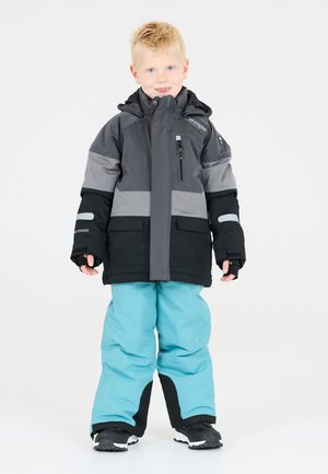 Giacca con cappuccio grigia e nera con tasca con zip; abbinata a pantaloni da neve blu chiaro e dettagli neri. Stivali da neve neri completano l'outfit.