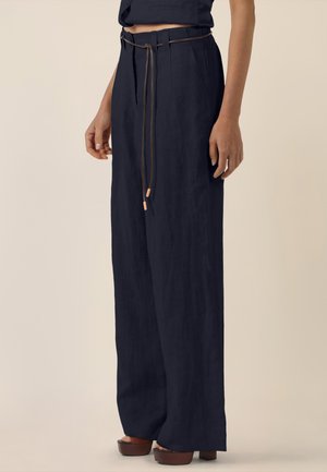 Frau trägt hoch taillierte, weit geschnittene marineblaue Hose mit dünnem Gürtel und Plateausandalen, steht vor einem schlichten beigen Hintergrund.