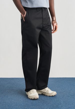 Fekete, egyenes szárú nadrág, sima anyagból, oldalzsebekkel és cipzáras sliccel. Világos színű tornacipővel párosítva.