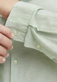 Camisa verde claro con una textura suave, que presenta un cierre de botón en el puño y detalles de costura sutiles.