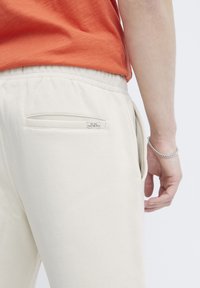 Pantalones de chándal beige con una textura suave, que cuentan con un bolsillo trasero y una pequeña etiqueta que dice "Eventos todo el día, todos los días". Cintura elástica visible.