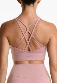Roze sportbeha met kruislings straps, zachte textuur en een aansluitende pasvorm; heeft een logo op de onderband. Gemaakt in combinatie met bijpassende hooggesneden legging.