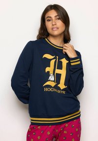 Donna che indossa una felpa blu navy con una grande "H" gialla e un'immagine di un gufo sopra la scritta "Hogwarts", abbinata a pantaloni rossi con disegni di logo "HP".