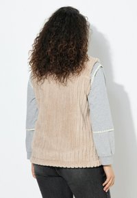 Gilet en polaire beige à côtes, avec une texture douce, superposé sur un haut à manches longues gris avec des accents au niveau des poignets. Vue de l'arrière.