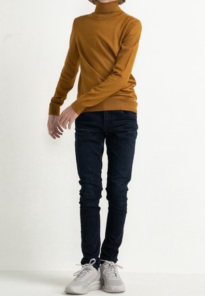 Personne portant un pull à col roulé moutarde, un jean skinny bleu foncé et des baskets blanches, debout devant un fond blanc uni.