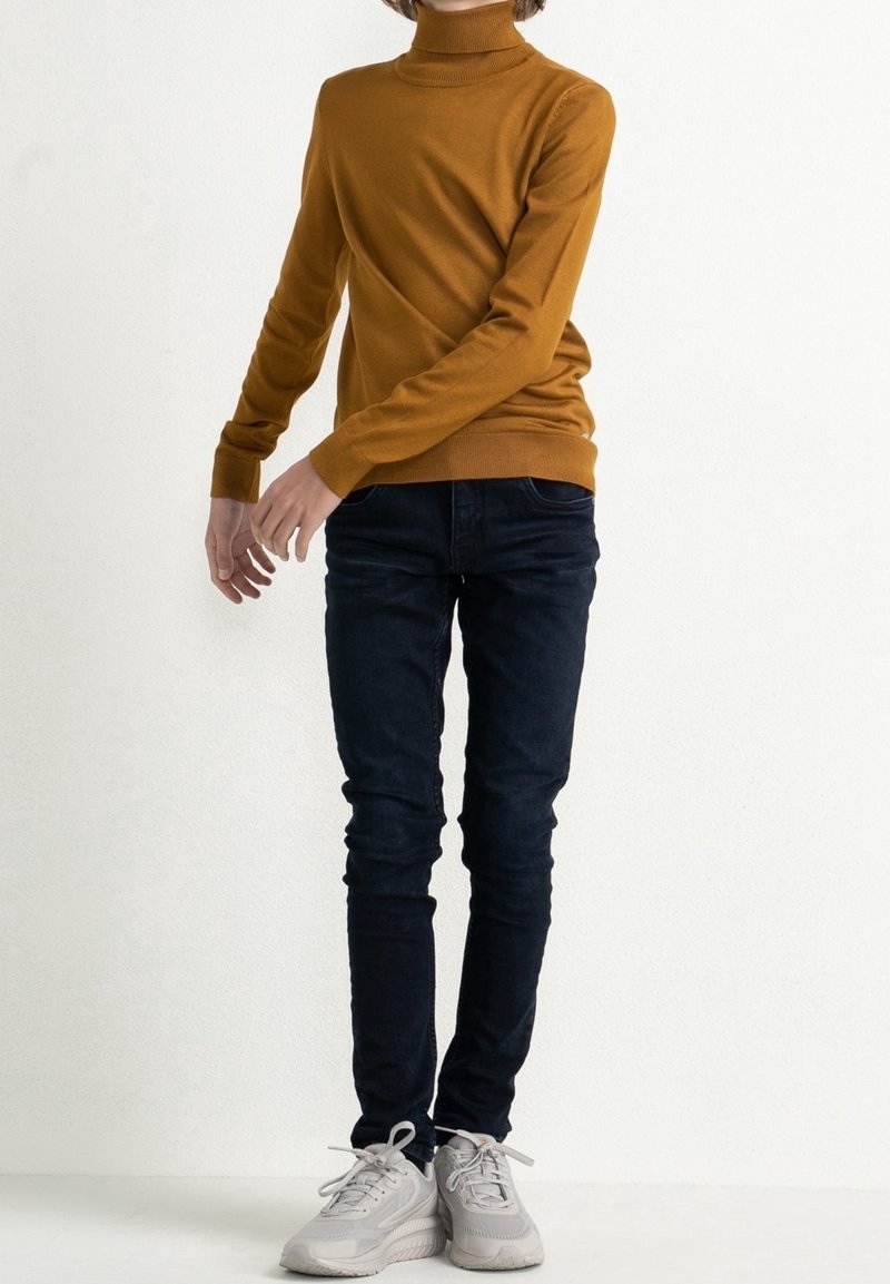 Personne portant un pull à col roulé moutarde, un jean skinny bleu foncé et des baskets blanches, debout devant un fond blanc uni.