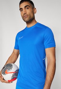 Blå idretts skjorte laget av glatt stoff, med en hvit Nike-logo og nettingpaneler på sidene; person som holder en mønstrete fotball.
