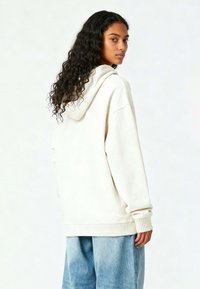 Felpa color crema realizzata in morbido tessuto con un ampio cappuccio, vestibilità rilassata, polsini a costine e un design semplice, abbinata a pantaloni larghi di un blu chiaro.