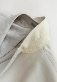 Indumento beige chiaro con uno strato esterno testurizzato e una fodera interna liscia. Presenta un colletto di colore chiaro in contrasto e dettagli di cucitura precisi.