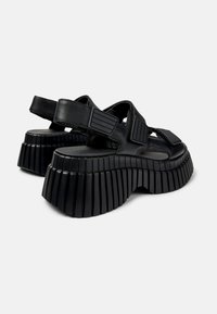 Black platform sandals with a thick, ribbed sole and textured straps featuring rectangular accents. Made of leather with open sides.