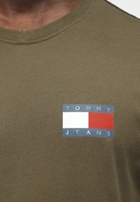 Κοντινή λήψη πουκάμισου ελιάς με ορθογώνιο λογότυπο Tommy Jeans σε μπλε, άσπρο και κόκκινο στο στήθος.