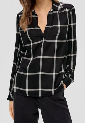 Kobieta ubrana w czarną bluzkę z długim rękawem z dużym białym wzorem w kratę i dekoltem w serek, w połączeniu z czarnymi spodniami, na jednolitym tle.