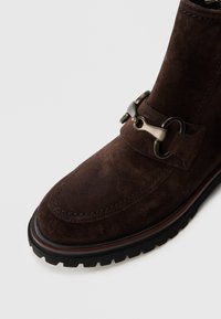 Bottine en suède brun foncé avec une épaisse semelle noire et un détail de boucle en métal décorative sur la section avant supérieure.
