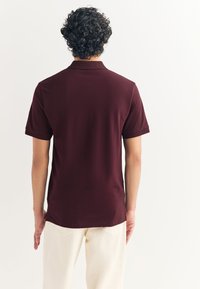 Bordó polokošile s krátkými rukávy, límečkem a hladkou texturou, nošená s krémovými kalhotami. Viditelné švy a detaily na míru.