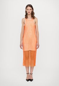 Robe en maille orange avec un design sans manches, longueur genoux, présentant des motifs en forme de losange et une texture semi-transparente. Escarpins noirs portés.