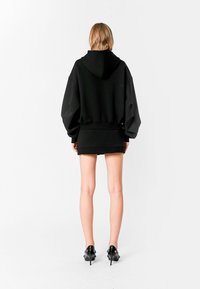 Sudadera negra de gran tamaño con capucha, mangas largas y un diseño corto, combinada con una falda mini negra y unos zapatos de tacón alto negros y brillantes.