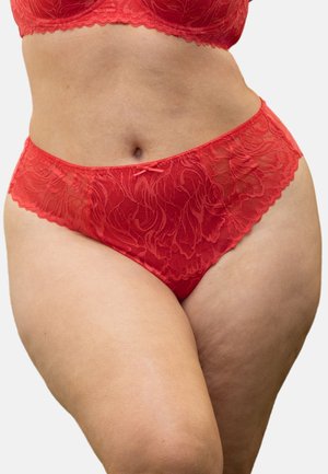 Frau trägt roten Spitzen-BH und passende hoch taillierte Unterhose und zeigt ihren Bauch und ihre Oberschenkel vor einem einfarbigen Hintergrund.