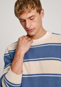 Blue ja kreemikad triibulised kudumipullid ribidega kaela- ja varrukatelt. Pehme tekstuur; sisaldab õhukesi valgeid aktsenttriipse.