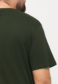 Tamnozelena majica s kratkim rukavima i okruglim izrezom; glatka tekstura, jednostavan dizajn, bez vidljivih uzoraka ili detalja.