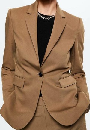 Person wearing a tailored tan blazer with black button, matching pants, black shirt, and a silver chain necklace.