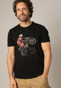 Man in een zwart T-shirt met een afbeelding van een oudere vrouw die op een motorfiets rijdt en een wheelie maakt, met de tekst "Last Life Deluxe."