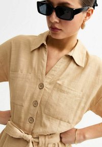 Robe chemise beige à manches courtes, fermeture boutonnée sur le devant, deux poches poitrine et ceinture à la taille. Accessoirisée avec des lunettes de soleil noires et des boucles d'oreilles créoles.