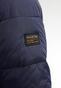 Chaqueta acolchada azul marino con una textura suave y brillante, con una etiqueta negra que dice "Petrol Ind" y detalles relacionados en naranja.