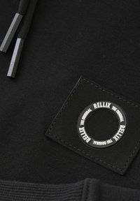 Zwarte stof met een textuur, rubberen logopatch met een cirkelvormig ontwerp en geperst tekst. De trekkoorden hebben metalen uiteinden.