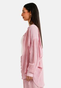 Camisa de color rosa claro, con pliegues y un corte holgado, de mangas largas. Presenta un cierre de botones y una textura suave, llevada sobre una blusa blanca.