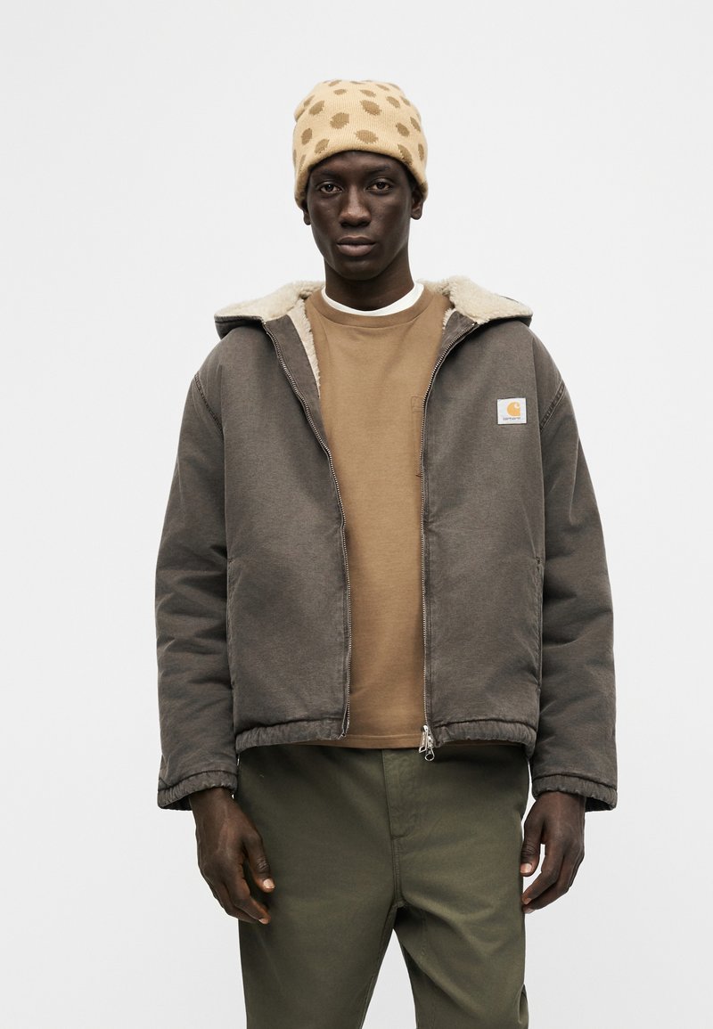 Mann trägt eine beige Mütze mit Polka-Dots, braune Fleecejacke mit Reißverschluss, ein beige Sweatshirt und olivgrüne Hosen, steht vor einem schlichten Hintergrund.