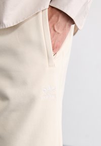 Cream-colored sweatpants made of soft fabric, featuring a side pocket and a white embroidered logo on the thigh. Minimalist design.