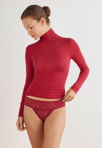 Haut à col roulé rouge à manches longues avec une texture lisse, assorti à une culotte en dentelle rouge ornée de motifs floraux et de bords festonnés.