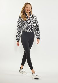 Schwarz-weißer Pullover mit abstraktem Muster und Reißverschlusskragen, kombiniert mit hochtaillierten schwarzen Leggings und weißen Sportschuhen.