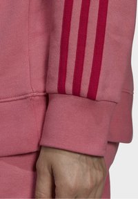 Detalj av rosa sweatshirt som visar en mudd med ribbad struktur och tre röda ränder längs ärmen. Mjukt bomullsblandat tyg.