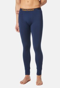 Männlicher Model trägt navyblaue, eng anliegende lange Unterwäsche-Leggings mit orangem "bruno banani"-Bund und steht barfuß vor weißem Hintergrund.