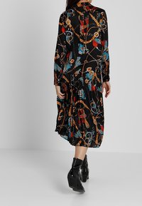 Černé šaty s dlouhými rukávy, s barevným vzorem řetězů a předmětů. Detail s plisovanou sukní. Nositelná s černými vysokými botami na podpatku.