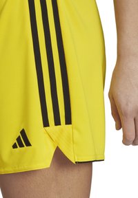 Gelbe Sportshorts aus glattem Material, mit schwarzen Seitenstreifen, einem schwarzen Logo und einem gemusterten Saumdetail am Beinabschluss.