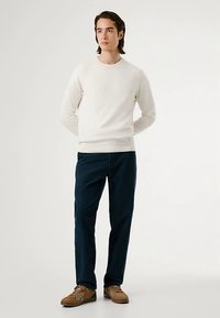 Kremfarget strikkegenser med rund hals, kombinert med mørkeblå bukser og brune sko. Antrekket er minimalistisk og uformelt.