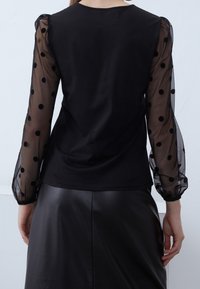 Schwarzes Oberteil mit transparenten Puffärmeln im Polka-Dot-Muster und einem glatten Oberteil, kombiniert mit einem schwarzen Kunstlederrock.