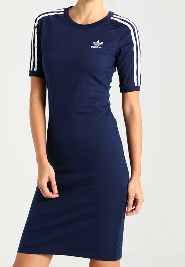 Femme portant une robe ajustée Adidas bleu marine avec des rayures blanches sur les manches et le logo Adidas sur la poitrine, se tenant devant un fond uni.